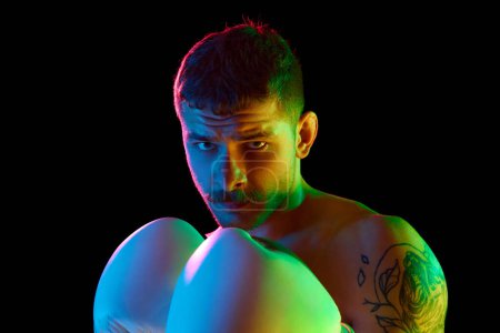 Photo for Portrait of concentrated, serious, brutal young man, boxing athlete standing shirtless with gloves isolated over black background in neon light. Concept of sport, combat sport, martial arts, strength - Royalty Free Image