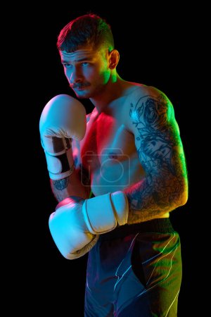 Photo for Portrait of concentrated young man, boxing athlete standing shirtless with gloves isolated over black background in neon light. Concept of professional sport, combat sport, martial arts, strength - Royalty Free Image