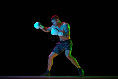 Téléchargez les photos : Jeune homme torse nu, musclé, boxeur en mouvement, s'entraînant isolé sur fond noir sous la lumière du néon. Concept de sport professionnel, sport de combat, arts martiaux, force - en image libre de droit
