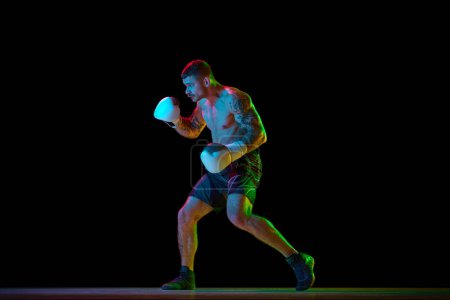 Photo for Full-length image of young athletic man, muscular, shirtless boxer in motion, fighting, practicing hooks isolated over black background in neon. Concept of sport, combat sport, martial arts, strength - Royalty Free Image