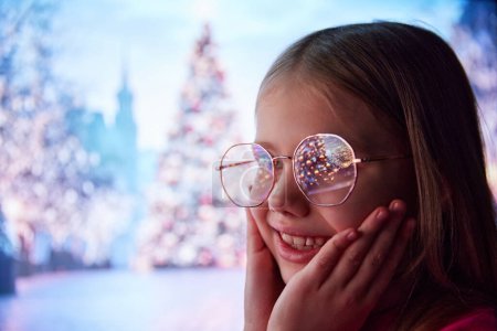 Téléchargez les photos : Petite fille, enfant debout à l'extérieur avec un visage souriant heureux. Arbre de Noël avec des lumières réfléchissant sur les lunettes pour enfants. Concept de Noël, enfance, rêves, fantaisie, bonheur, inspiration - en image libre de droit