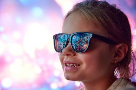Foto de Feliz niña sonriente, niña alegre en gafas de sol mirando fuegos artificiales. Reflexión sobre las gafas. Celebración al aire libre. Concepto de Navidad, infancia, sueños, fantasía, felicidad, diversión - Imagen libre de derechos