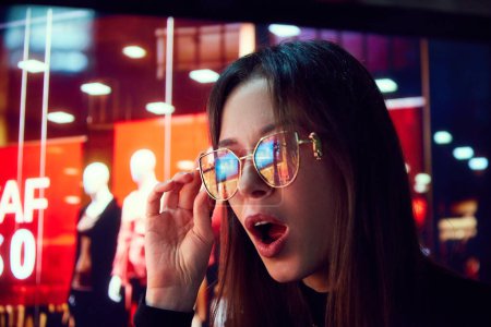 Foto de Mujer joven conmocionada y emocionada mirando en los escaparates con luces de neón reflejo en las gafas. Shopaholic, ventas. Concepto de emoción humana, compras, ventas, vacaciones - Imagen libre de derechos