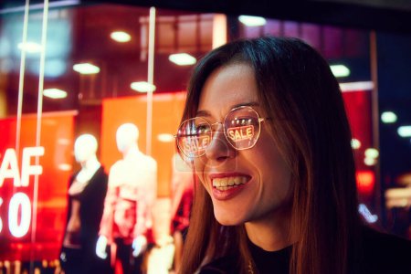 Photo for Close-up portrait of smiling young woman with reflection on glasses looking on shop windows. Big sales in shopping mall. Concept of human emotion, shopping, sales, holidays - Royalty Free Image