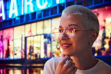 Foto de Joven mujer sonriente de pie en la calle de la noche y mirando en las ventanas del centro comercial. Luces de reflexión sobre las gafas. Concepto de compras, temporada de ventas, emociones humanas, vacaciones - Imagen libre de derechos