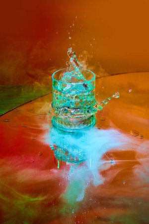 Photo for Ice cube falling down into glass with alcohol against orange background in neon light with smoke effect. Concept of alcohol drink, nightclub, party, taste, celebration. - Royalty Free Image