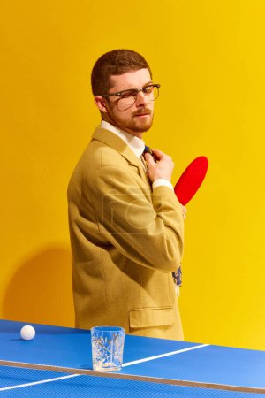 Foto de Hombre joven guapo en ropa elegante jugando al tenis tableta, mirando en vaso de whisky vacío sobre fondo amarillo brillante. Ganador. Concepto de deporte, ocio, hobby, creatividad. Arte pop - Imagen libre de derechos