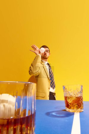 Téléchargez les photos : Jeune homme, homme d'affaires en costume élégant payer ping-pong, visant balle dans le verre de whisky sur fond jaune vif. Concept de sport, de loisirs, de passe-temps, de créativité, de plaisir et de joie. Pop art - en image libre de droit