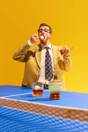 Téléchargez les photos : Ping-pong. Homme d'affaires en costume élégant buvant du whisky et snacking balle de tennis sur fond jaune vif. Concept de sport, de loisirs, de passe-temps, de créativité, de plaisir et de joie. Pop art - en image libre de droit