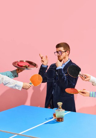 Téléchargez les photos : Homme réfléchi en costume élégant choisissant des raquettes de tennis de table sur fond rose. Photographie créative. Concept de sport, de loisirs, de passe-temps, de créativité, de plaisir et de joie. Pop art - en image libre de droit