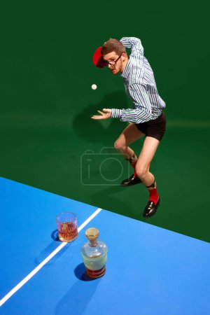 Téléchargez les photos : Vue de dessus. Jeune homme concentré en chemise et sous-vêtements jouant au tennis de table avec du verre de whisky sur fond vert. Concept de sport, de loisirs, de passe-temps, de créativité, de plaisir et de joie. Pop art - en image libre de droit