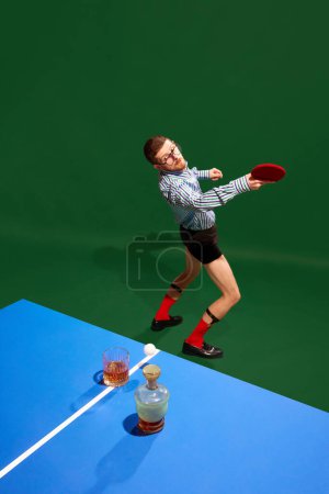 Téléchargez les photos : Vue de dessus. Jeune homme concentré en chemise et sous-vêtements jouant au tennis de table avec du verre de whisky sur fond vert. Concept de sport, de loisirs, de passe-temps, de créativité, de plaisir et de joie. Pop art - en image libre de droit