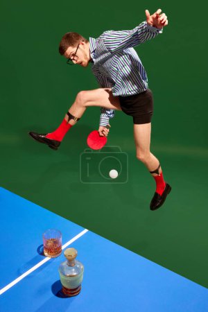 Téléchargez les photos : Vue de dessus. Jeune homme en chemise et sous-vêtements en mouvement. sauter, jouer au tennis sur tablette, boire du whisky sur fond vert. Concept de sport, de loisirs, de passe-temps, de créativité, de plaisir et de joie. Pop art - en image libre de droit