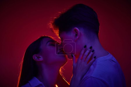 Foto de Precioso joven hombre y mujer besándose tiernamente, mostrando amor y cuidado contra el fondo rojo en luz de neón. Concepto de romance, amor, relación, pasión, juventud, citas, felicidad - Imagen libre de derechos