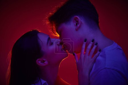 Foto de Retrato de joven hombre y mujer, pareja cariñosa besándose sobre fondo rojo en luz de neón. Aniversario. Concepto de romance, amor, relación, pasión, juventud, citas, felicidad - Imagen libre de derechos