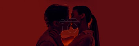 Foto de Apasionado joven hombre y mujer, joven pareja besándose, expresando amor contra fondo rojo en luz de neón. Monocromo. Concepto de romance, amor, relación, pasión, juventud. Banner - Imagen libre de derechos