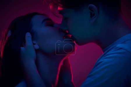 Foto de Mujer joven tierna besando a su novio, mostrando amor y cuidado contra el fondo púrpura en luz de neón. Concepto de romance, amor, relación, pasión, juventud, citas, felicidad - Imagen libre de derechos