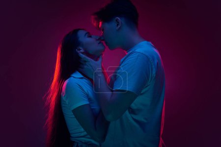 Foto de Novio y novia, joven hombre y mujer besándose contra fondo púrpura en luz de neón. Concepto de romance, amor, relación, pasión, juventud, citas, felicidad - Imagen libre de derechos