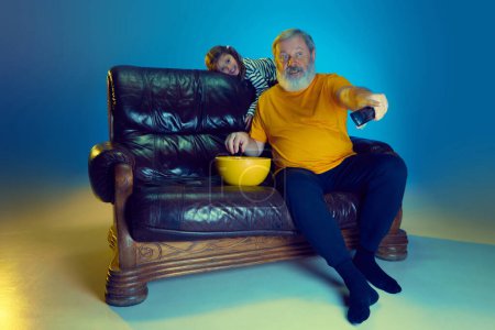 Photo for Happy senior man, grandfather sitting on couch at home with his little granddaughter and watching TV over blue background in neon light. Concept of family, happiness, care and love, emotions, leisure - Royalty Free Image