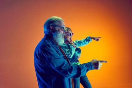 Téléchargez les photos : Homme âgé excité et émotionnel, et petite fille regardant un film en lunettes 3D sur fond orange dégradé sous la lumière du néon. Concept de famille, bonheur, soins et amour, unité, émotions, loisirs - en image libre de droit