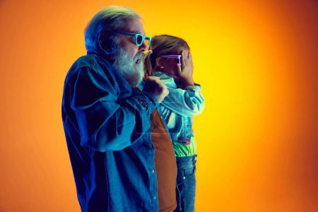Téléchargez les photos : Un film d'horreur. Homme âgé regardant un film avec une petite fille sur fond orange dégradé dans la lumière du néon. Concept de famille, bonheur, soins et amour, unité, émotions, loisirs - en image libre de droit
