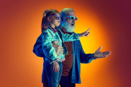 Téléchargez les photos : Grand-père regardant un film avec sa petite-fille sur fond orange dégradé sous la lumière du néon. Concept de famille, bonheur, soins et amour, unité, émotions, loisirs - en image libre de droit