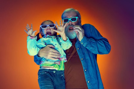 Photo for Horror movie. Senior man watching movie with little girl over gradient orange background in neon light. Concept of family, happiness, care and love, unity, emotions, leisure - Royalty Free Image