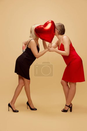 Foto de Dos chicas, pareja lesbiana en vestidos besándose detrás de globo de aire en forma de corazón sobre fondo beige claro. Concepto de comunidad lgbt, amor, día de San Valentín, libertad, aceptación, 14 de febrero - Imagen libre de derechos