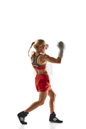 Téléchargez les photos : Athlète de boxe musclée et compétitive, jeune femme en mouvement, pratiquant, frappant isolée sur fond blanc. Concept de sport, mode de vie actif et sain, force, endurance, soins du corps - en image libre de droit