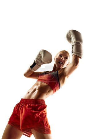 Téléchargez les photos : Athlète de boxe musclée et compétitive, jeune femme en mouvement, pratiquant, frappant isolée sur fond blanc. Concept de sport, mode de vie actif et sain, force, endurance, soins du corps - en image libre de droit