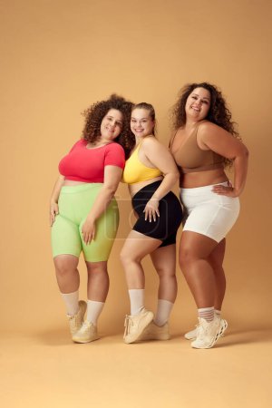 Full-length image of three attractive young women, friends with fat, overweigh bodies standing in sportswear over beige background. Concept of sport, body-positivity, weight loss, body and health care