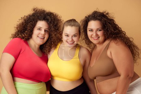 Photo for Portrait of three beautiful, smiling, overweigh women standing in sportswear over beige studio background. Concept of sport, body-positivity, weight loss, body and health care - Royalty Free Image