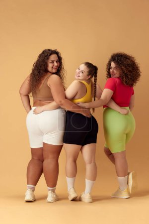 Photo for Back view of three smiling young omen, plus size models in comfortable sportswear over beige studio background. Concept of sport, body-positivity, weight loss, body and health care - Royalty Free Image