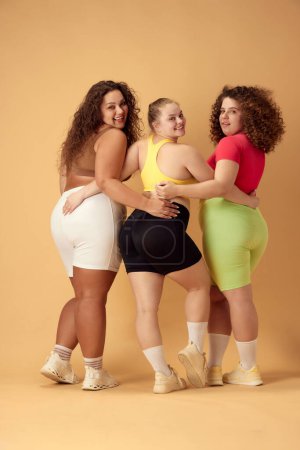 Foto de La imagen completa de tres mujeres jóvenes atractivas, amigas con grasa, sobrepesan los cuerpos de pie en ropa deportiva sobre un fondo beige. Concepto de deporte, positividad corporal, pérdida de peso, cuidado corporal y de la salud - Imagen libre de derechos