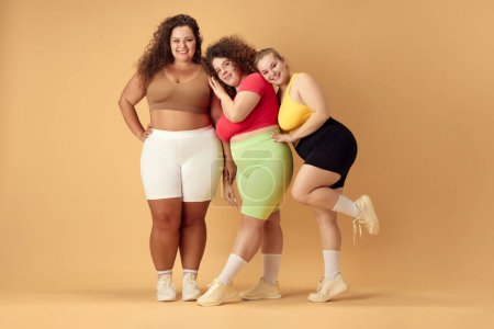 Photo for Full-length image of three attractive young women, friends with fat, overweigh bodies standing in sportswear over beige background. Concept of sport, body-positivity, weight loss, body and health care - Royalty Free Image