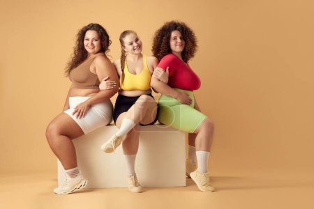 Téléchargez les photos : Trois belles jeunes femmes, plus des modèles de taille avec des corps surdimensionnés, posant en vêtements de sport sur fond de studio beige. Concept de sport, positivité corporelle, perte de poids, corps et soins de santé - en image libre de droit