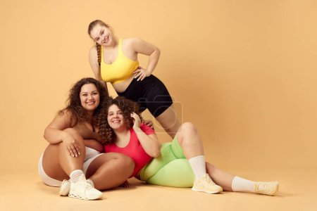 Photo for Three beautiful young women, plus size models with oversized bodies, posing in sportswear over beige studio background. Concept of sport, body-positivity, weight loss, body and health care - Royalty Free Image