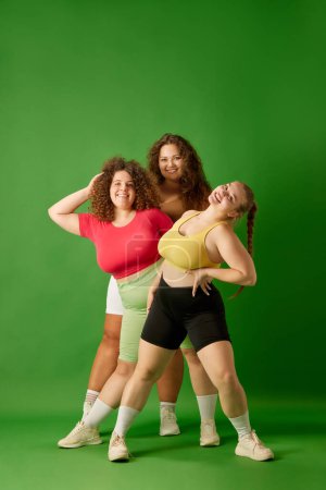 Foto de Sonrientes, hermosas mujeres jóvenes con formas de cuerpo de gran tamaño, en ropa deportiva posando sobre fondo de estudio verde. Concepto de deporte, positividad corporal, pérdida de peso, cuidado corporal y de la salud - Imagen libre de derechos