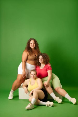 Foto de Sonrientes, hermosas mujeres jóvenes con formas de cuerpo de gran tamaño, en ropa deportiva posando sobre fondo de estudio verde. Concepto de deporte, positividad corporal, pérdida de peso, cuidado corporal y de la salud - Imagen libre de derechos