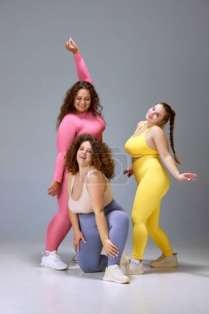 Foto de Aceptación. Tres mujeres jóvenes sonrientes en ropa deportiva cómoda de pie sobre fondo gris estudio. Además de belleza de tamaño. Concepto de deporte, positividad corporal, pérdida de peso, cuidado corporal y de la salud - Imagen libre de derechos