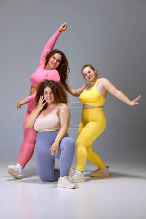 Photo for Acceptance. Three smiling young women in comfortable sportswear standing against grey studio background. Plus size beauty. Concept of sport, body-positivity, weight loss, body and health care - Royalty Free Image