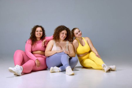 Photo for Support. Three beautiful women with oversized bodies, wearing sportswear, posing against grey studio background. Concept of sport, body-positivity, weight loss, body and health care - Royalty Free Image