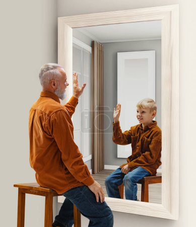 Photo for Senior man sitting near mirror and waving at his reflection as little boy. Pleasant memories of childhood. Conceptual collage. Concept of present, past and future, age, life cycle, generation, ad - Royalty Free Image