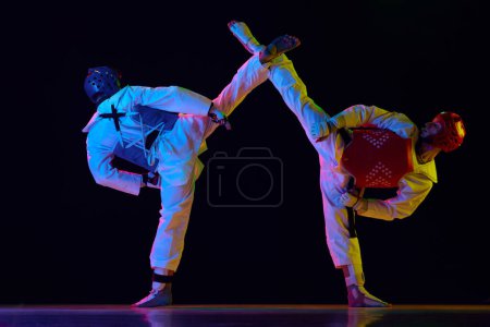 Téléchargez les photos : Deux hommes en kimono et casques pratiquant le taekwondo, s'entraînant, luttant contre un fond noir au néon. Concept d'arts martiaux, sport de combat, compétition, action, force - en image libre de droit
