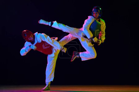 Photo for Dynamic image of young men, taekwondo athletes in kimono and helmets training against black background in neon light. Concept of martial arts, combat sport, competition, action, strength - Royalty Free Image