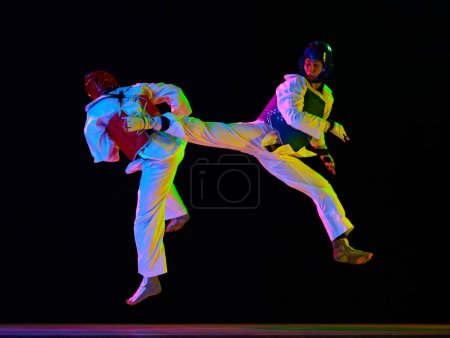 Foto de Imagen dinámica de hombres jóvenes, deportistas taekwondo en kimono y cascos entrenando sobre fondo negro en luz de neón. Concepto de artes marciales, deporte de combate, competencia, acción, fuerza - Imagen libre de derechos