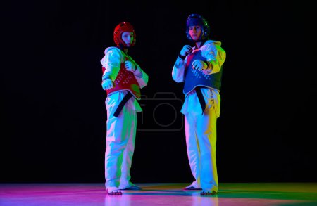 Photo for Two men, taekwondo athletes in white kimono and helmet standing, posing against black background in neon light. Concept of martial arts, combat sport, competition, action, strength - Royalty Free Image