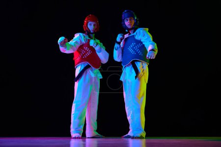 Téléchargez les photos : Deux hommes, athlètes de taekwondo en kimono blanc et casque debout, posant sur fond noir au néon. Concept d'arts martiaux, sport de combat, compétition, action, force - en image libre de droit