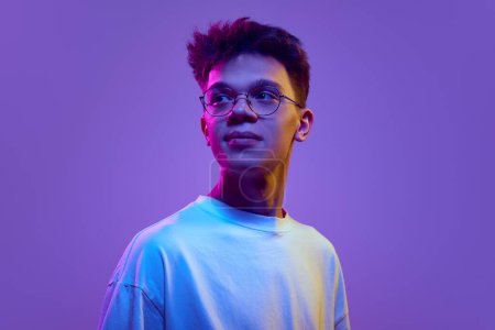 Photo for Portrait of smiling young man, boy in casual white t-shirt and sunglasses looking away against purple background in neon light. Relaxed look. Concept of human emotions, youth, education, lifestyle, ad - Royalty Free Image