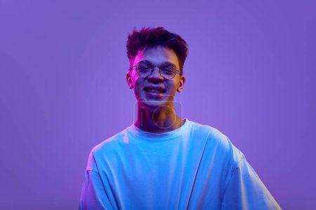 Photo for Portrait of happy smiling young man in casual clothes and glasses standing against purple background in neon light. Concept of human emotions, youth, education, positivity, education - Royalty Free Image